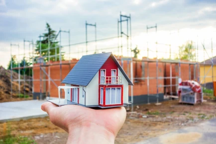 Baubegleitung nicht nur beim Neubau: Eine Hand hält ein Modellhaus, im Hintergrund ist eine Baustelle zu sehen